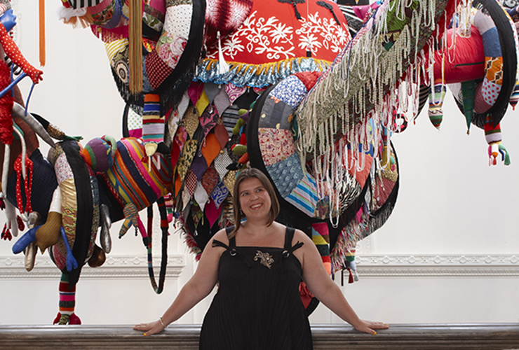 A light-skinned adult woman with bobbed brown hair wears a black sleeveless dress. She smiles wide and leans her hands back against a railing. Behind her is the lower portion of a large colorful fabric sculpture in front of a white wall.