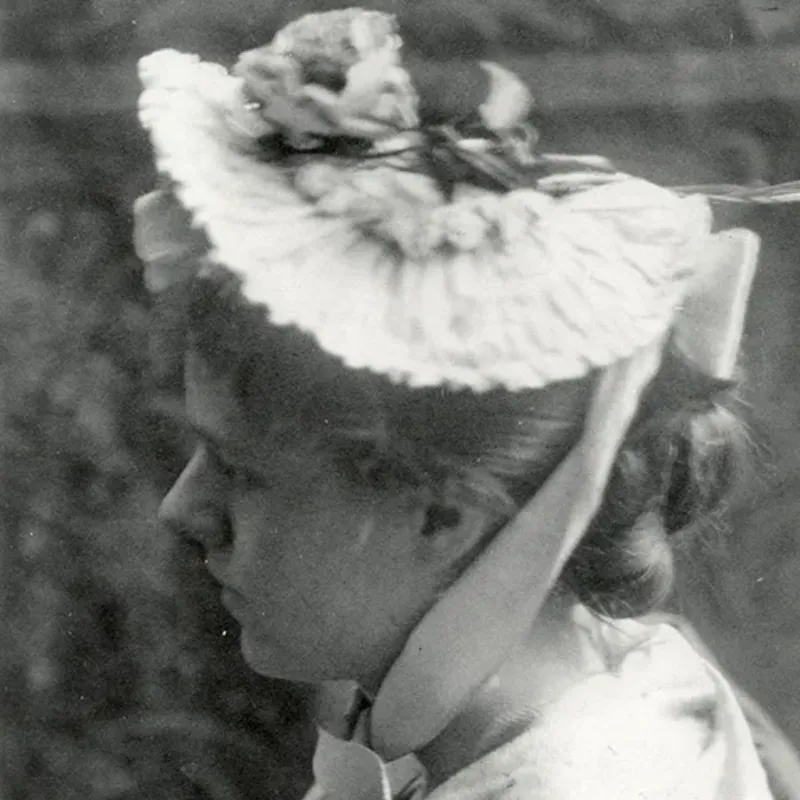 A soft-focused black-and-white photograph of a young light-skinned woman. She is in profile, and her light-colored hair is pulled into a bun. She wears a ruffled hat, with a sash tied under her chin. She is positioned in profile looking to the left of the image.