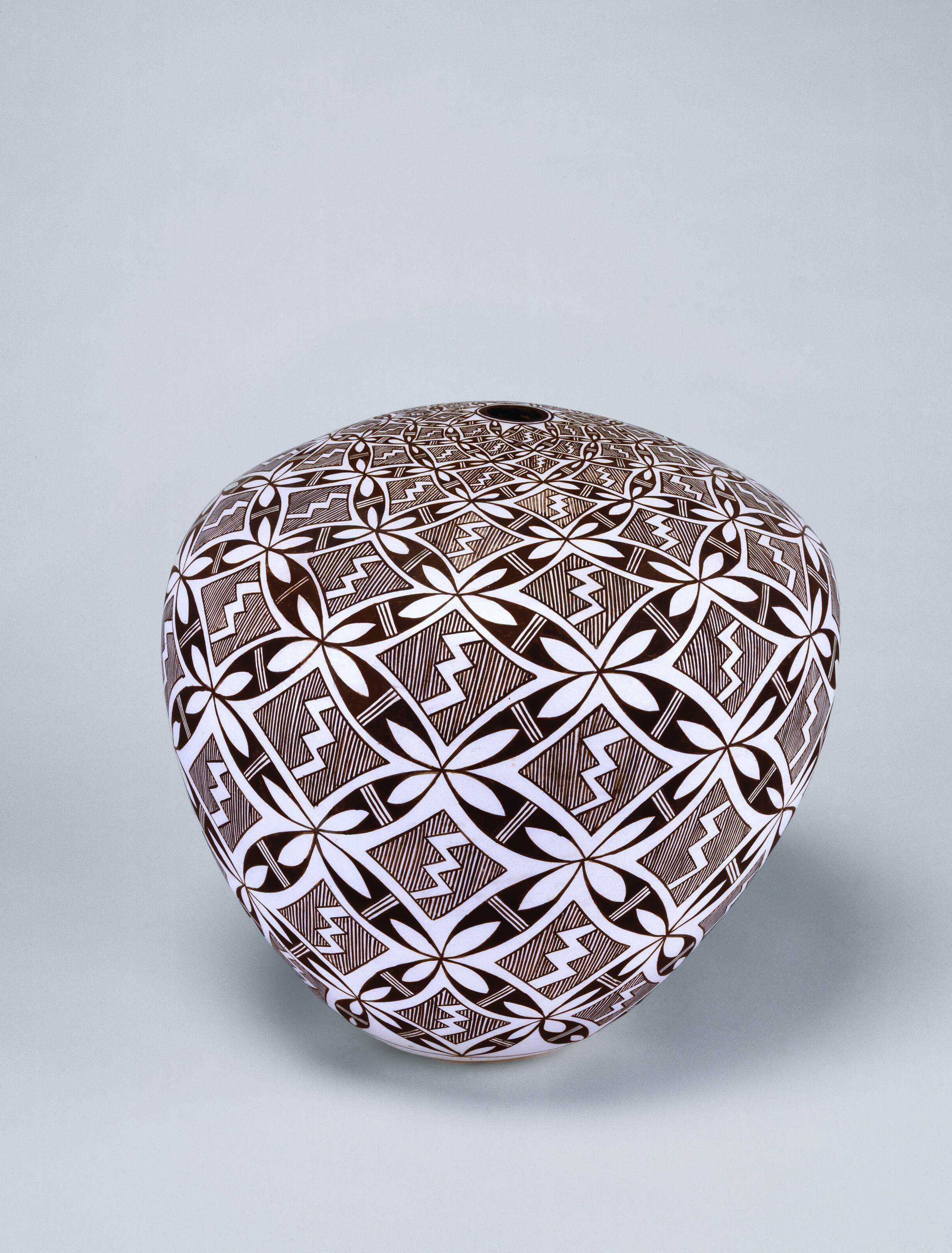 Round with a slight taper toward the base and a tiny opening at the top, this clay seed jar features a repeating pattern of abstracted flower shapes and a lightning bolt design. On the white surface the dark brown, delicately drawn designs expand and contract accordant with the form of the vessel.