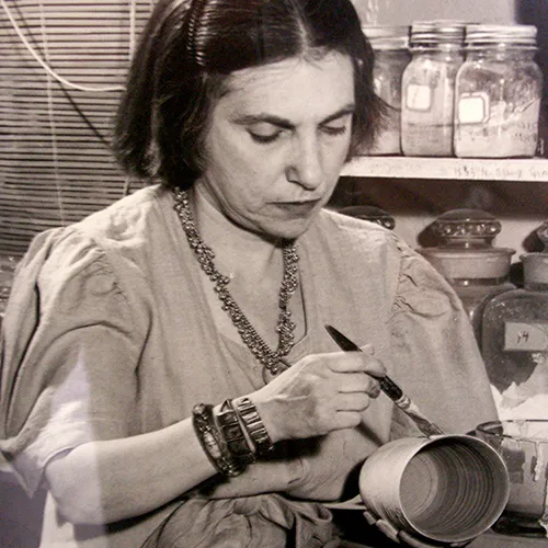 A black-and-white photograph of a light-skinned adult woman with short dark hair pinned away from her face. She brushes glaze onto a vase-shaped ceramic vessel in her other hand. She wears a smock-like dress, thick bracelets, and a necklace. Behind her are labeled mason jars.