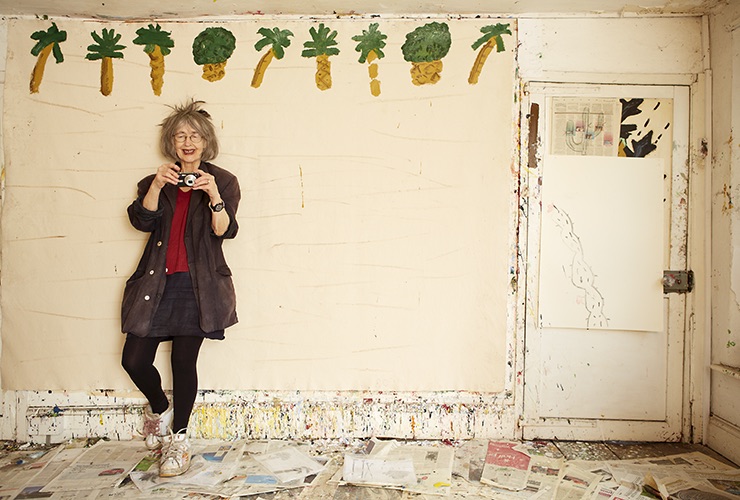 Color photograph of a light-skinned woman with grey hair holding a small digital camera. She stands in a delapidated room that has simple palm trees and pineapples painted at the top of one wall. Smiling, she wears white sneakers and props one foot up against the wall.