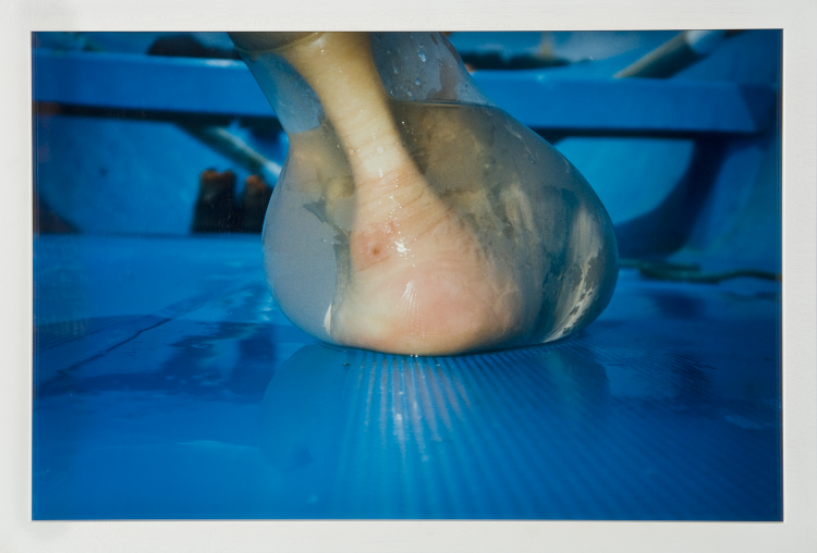A photograph shot at ground level captures an extreme close-up of a heel and ankle from behind. Enclosed in a water-filled latex balloon that distorts its shape, the foot rests upon a wet, sky blue surface that resembles the interior hull of a rowboat.
