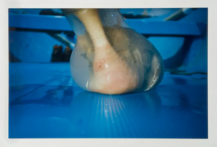 A photograph shot at ground level captures an extreme close-up of a heel and ankle from behind. Enclosed in a water-filled latex balloon that distorts its shape, the foot rests upon a wet, sky blue surface that resembles the interior hull of a rowboat.