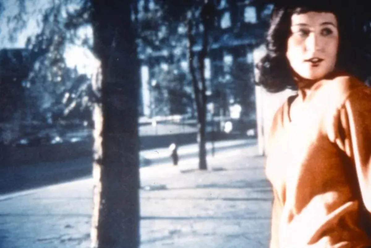 A woman stands in an orange sweater, her body turned to the center while she looks over her left shoulder, her mouth slightly open. Her dark hair is shoulder-length and contrasts with her light skin. Behind her, a screen with the image of trees and a building projected onto it.