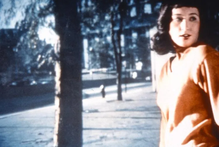 A woman stands in an orange sweater, her body turned to the center while she looks over her left shoulder, her mouth slightly open. Her dark hair is shoulder-length and contrasts with her light skin. Behind her, a screen with the image of trees and a building projected onto it.