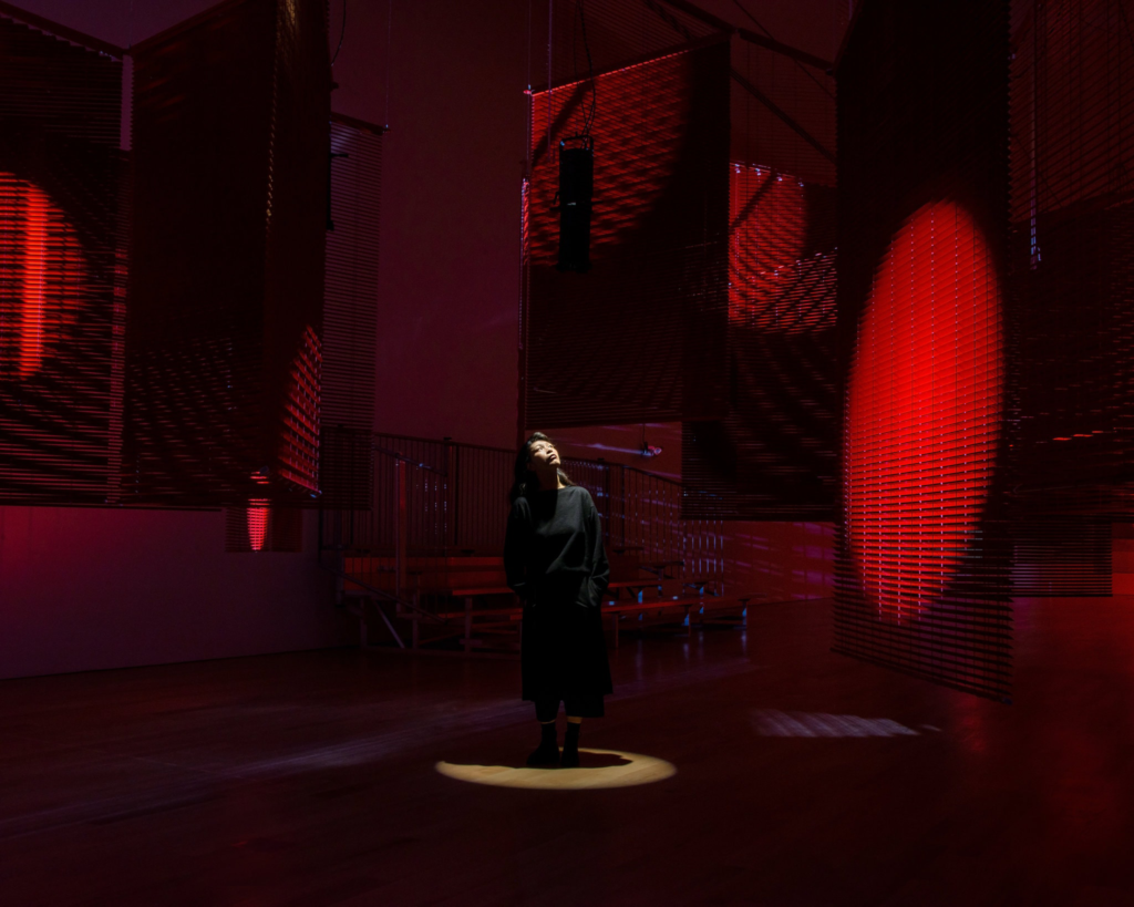 Artist Haegue Yang stands in a spotlight in the middle of her immersive exhibition, in a room with red lighting, and big banner-like structures hung from the ceiling. She looks up at one of the structures that has a red circle of light projected on it.