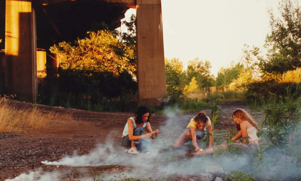 Crouching in the shadow of a highway overpass, three teenage girls with light skin focus intently on lighting smoke bombs.