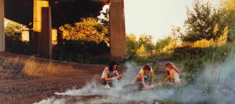 Crouching in the shadow of a highway overpass, three teenage girls with light skin focus intently on lighting smoke bombs.