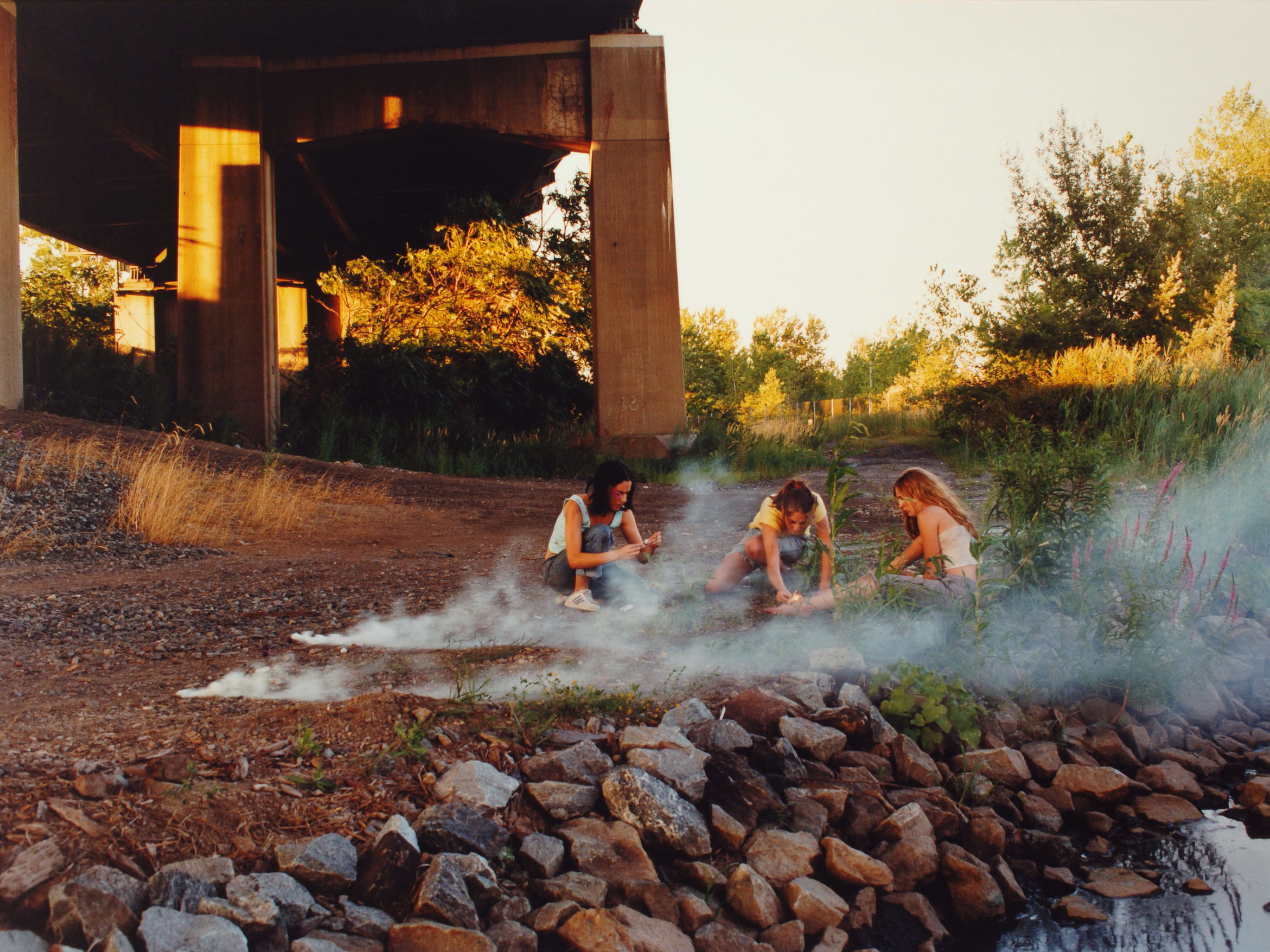 Crouching in the shadow of a highway overpass, three teenage girls with light skin focus intently on lighting smoke bombs.