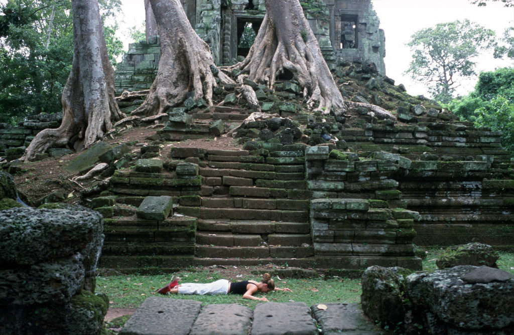 Photograph of ancient ruins with a woman lying facedown on grass in front of moss-covered stone steps that lead to a stone structure.