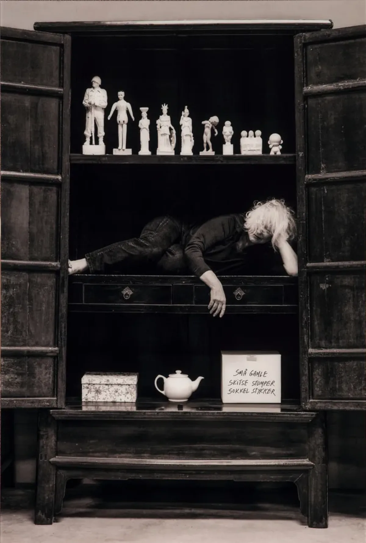 Black and white photograph of a woman with light skin and hair stuffed in a cabinet on her side with her head resting on her hand.