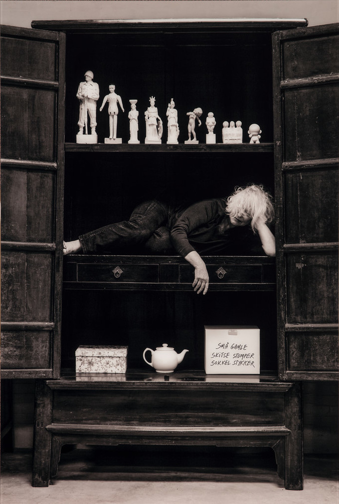 Black and white photograph of a woman with light skin and hair stuffed in a cabinet on her side with her head resting on her hand.