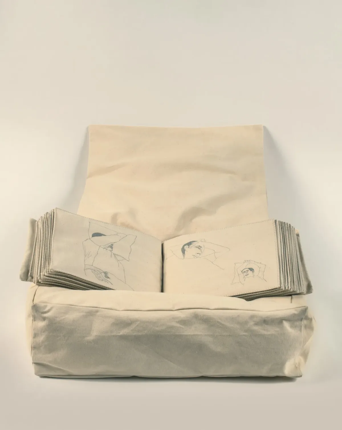 A stuffed, pillow-like box in beige holds an illustrated book with fabric pages. Three drawings of people sleeping fill the open book.