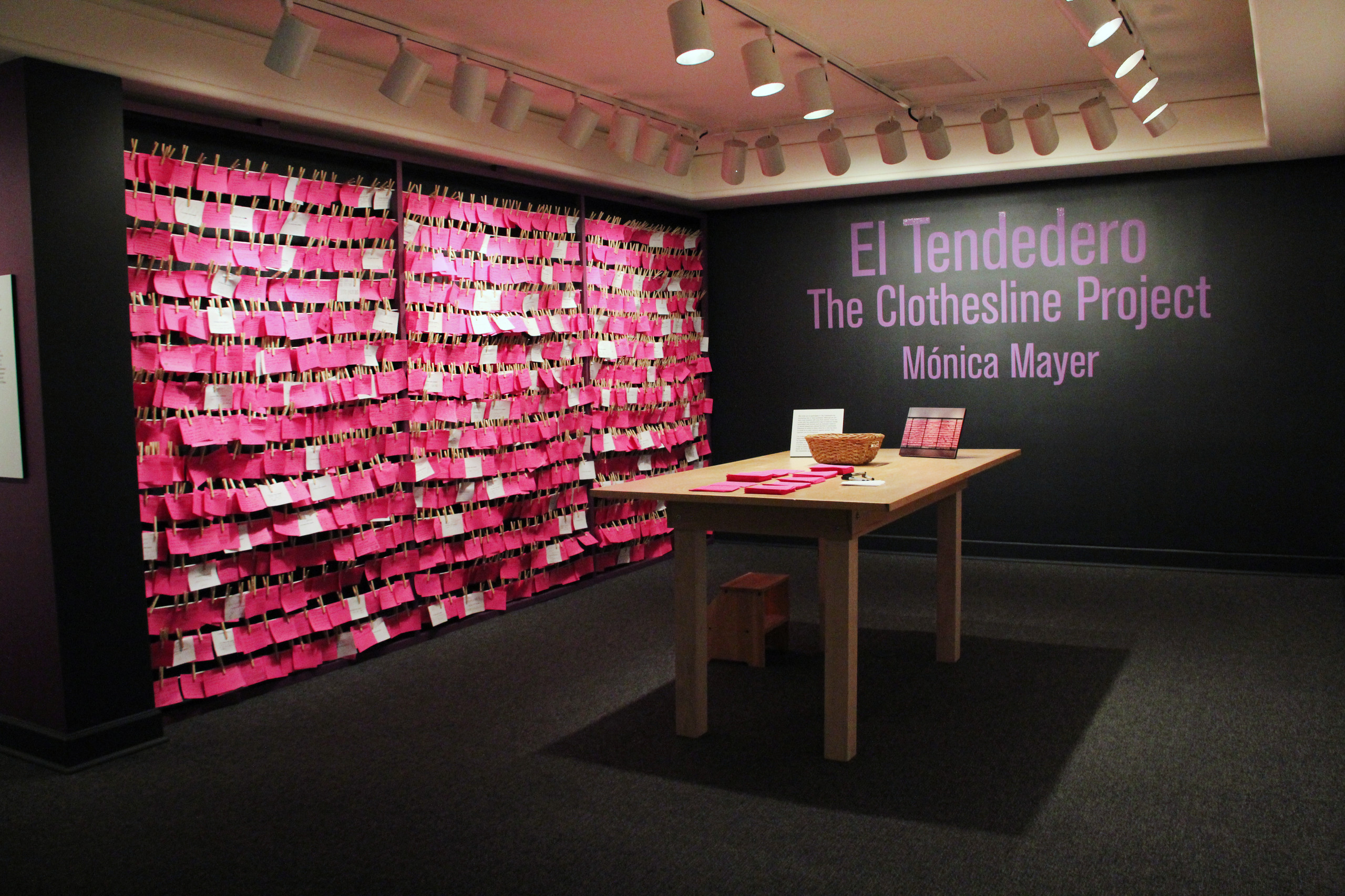 View of exhibition gallery features bright pink post-it notes line the wall from ceiling to floor. On the adjacent wall are the words in pink text: