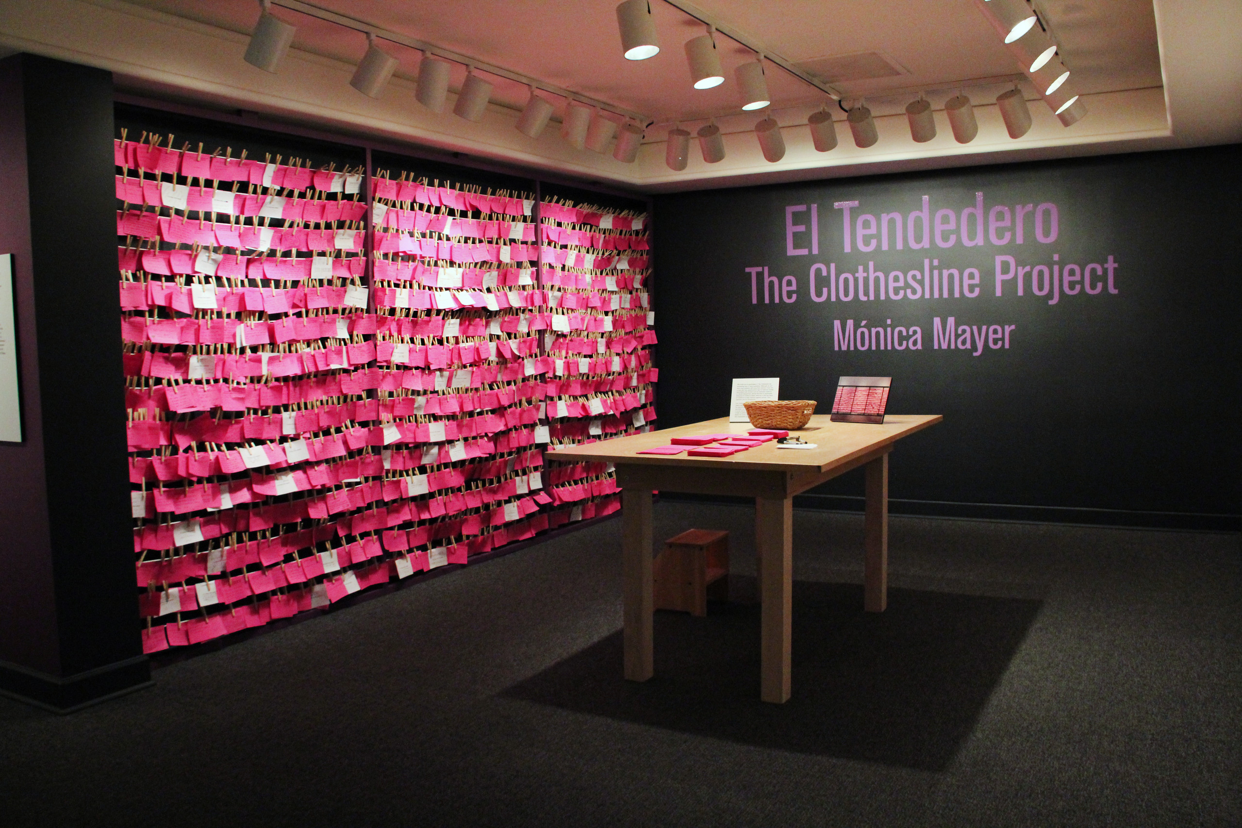 View of exhibition gallery features bright pink post-it notes line the wall from ceiling to floor. On the adjacent wall are the words in pink text:"El Tendedero, The Clothesline Project, Monica Meyer