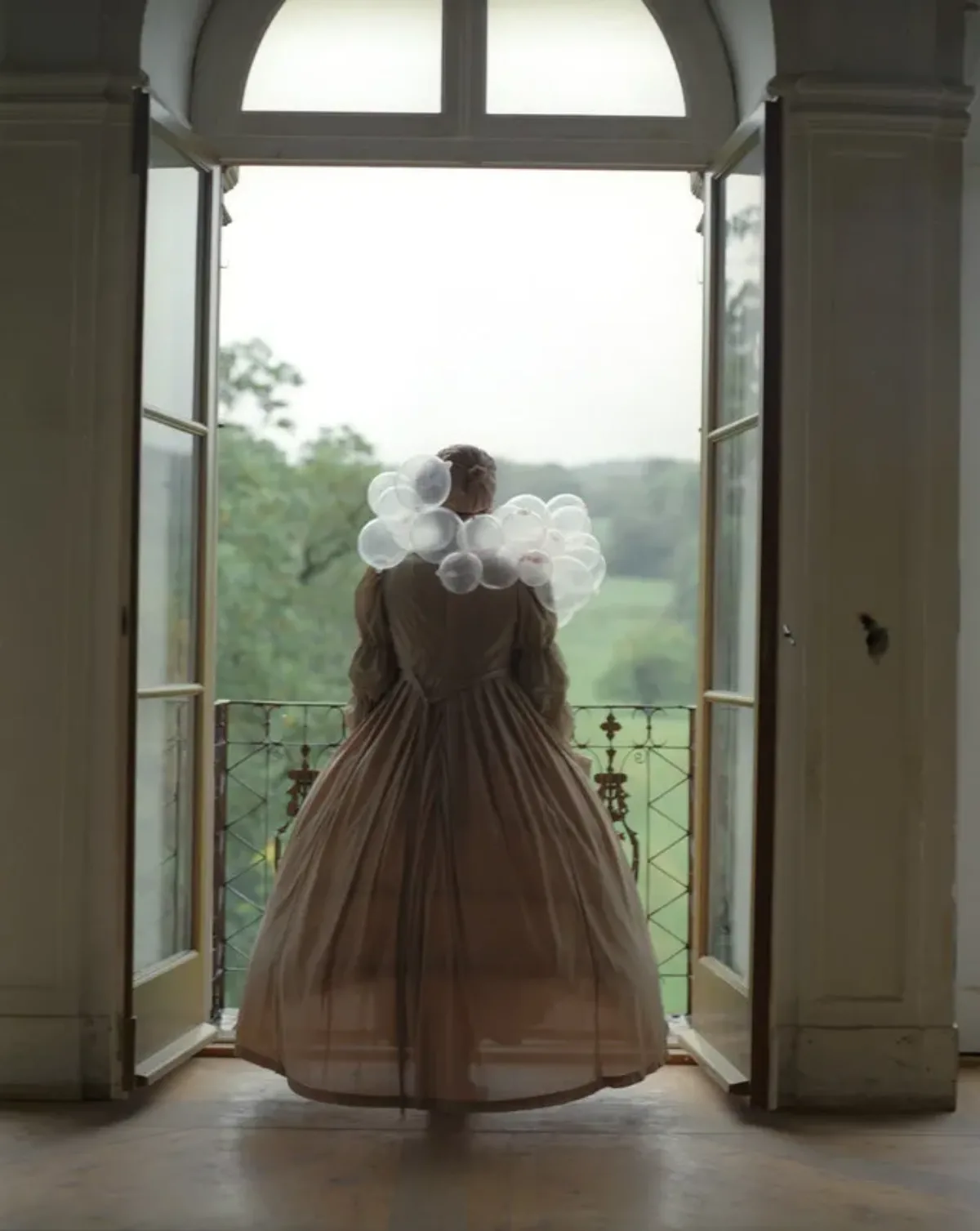 An adult woman seen from behind standing before two open glass doors leading out to a balcony overlooking greenery. She is dressed in a long, tan, period gown and her hair is tied up. Around her shoulders are several clear bubbles, resembling a scarf made of bubbles.