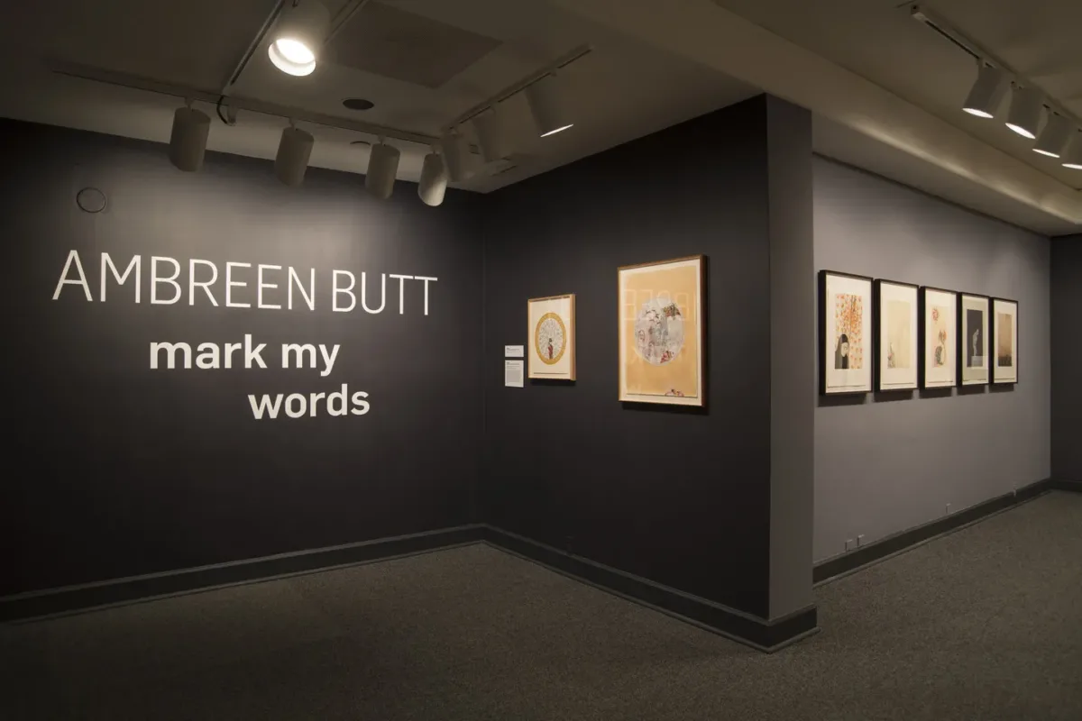 Installation view of seven works by Ambreen Butt on dark gray walls. The words "AMBREEN BUTT: mark my words" hand on the wall.