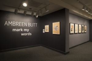 Installation view of seven works by Ambreen Butt on dark gray walls. The words "AMBREEN BUTT: mark my words" hand on the wall.
