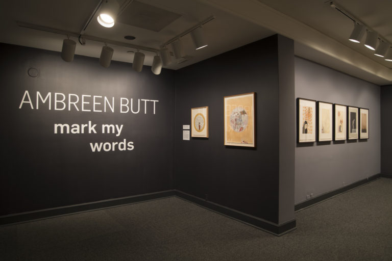Installation view of seven works by Ambreen Butt on dark gray walls. The words "AMBREEN BUTT: mark my words" hand on the wall.