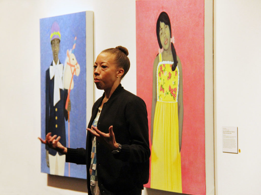 Amy Sherald speaks to a crowd in a gallery beside her striking portraits depicting individuals with gray skintones against vibrant, solid-colored backgrounds. She gestures with her hands and has a serious expression on her face.