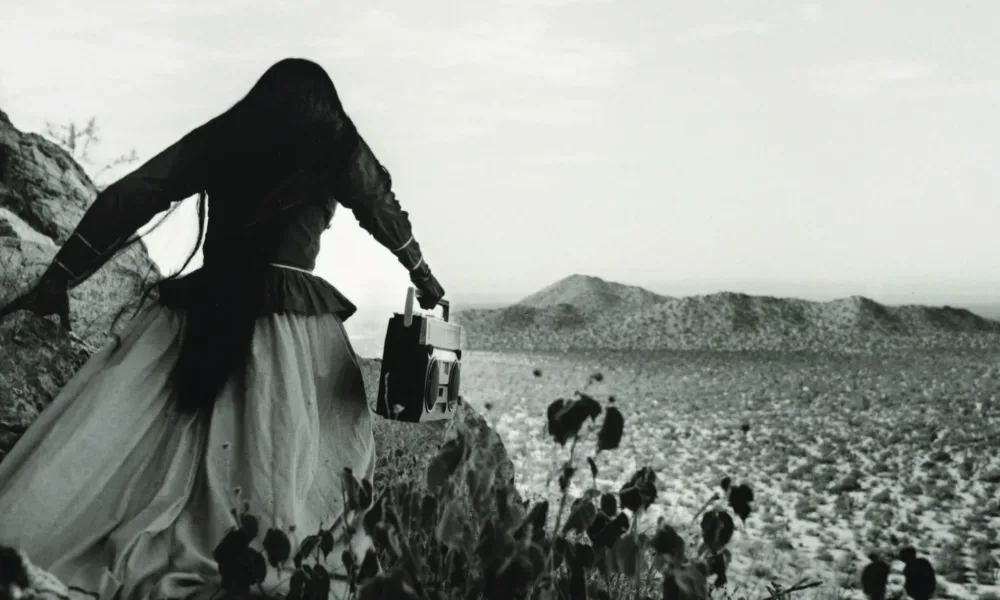 A black-and-white photograph shows the back of a woman as she crests a rocky path above a vast desert landscape beneath an expansive sky. Her traditional, ethnic full skirt, long-sleeved blouse, and long, straight, dark hair contrasts with the modern portable stereo she carries.