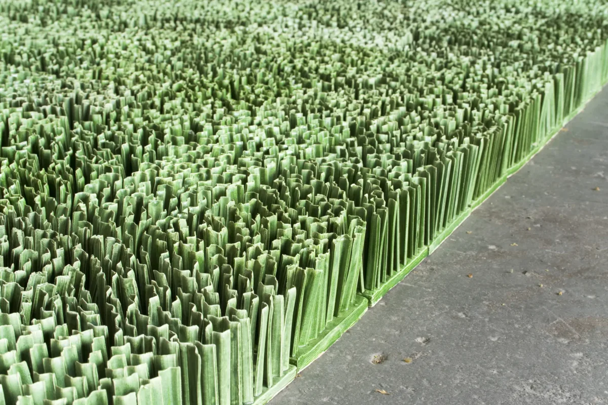 Detail photograph of ceramic sculpture made to look like a patch of lawn. Individual squares consisting of multiple upright blades of porcelain grass, glazed green, fit together to form a lush rectangular field of grass.