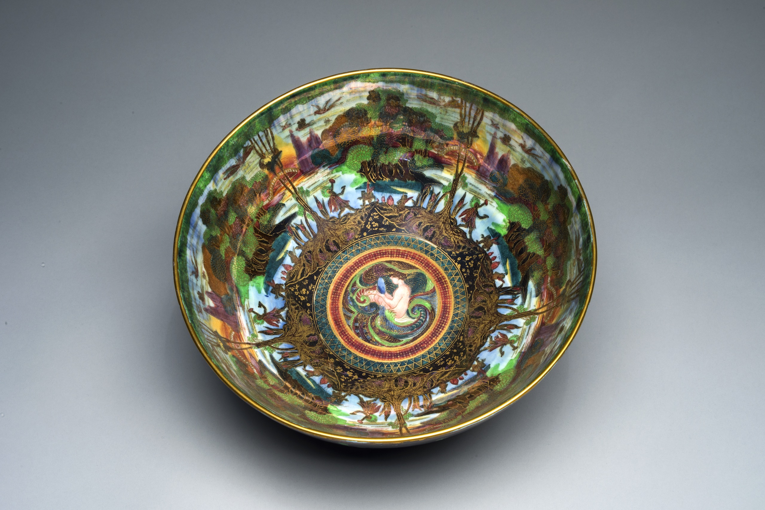 Interior view of a colorful, iridescent lusterware bowl featuring gold cloisonne and an ornamental Art Nouveau design. At the center, a light-skinned mermaid bathes in the ocean. Radiating outward, and repeated five times, is a lush agrarian scene.