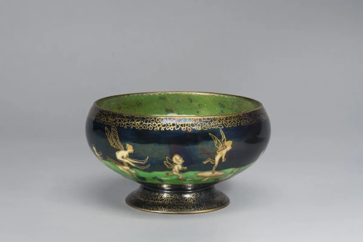 Porcelain pedestal bowl featuring an iridescent lustreware glaze. Around the bowl, winged fairies and sprites cavort on a vibrant green field set against a deep blue background. The figures and the decorative elements along the rim and foot of the bowl are outlined in gold cloisonne.