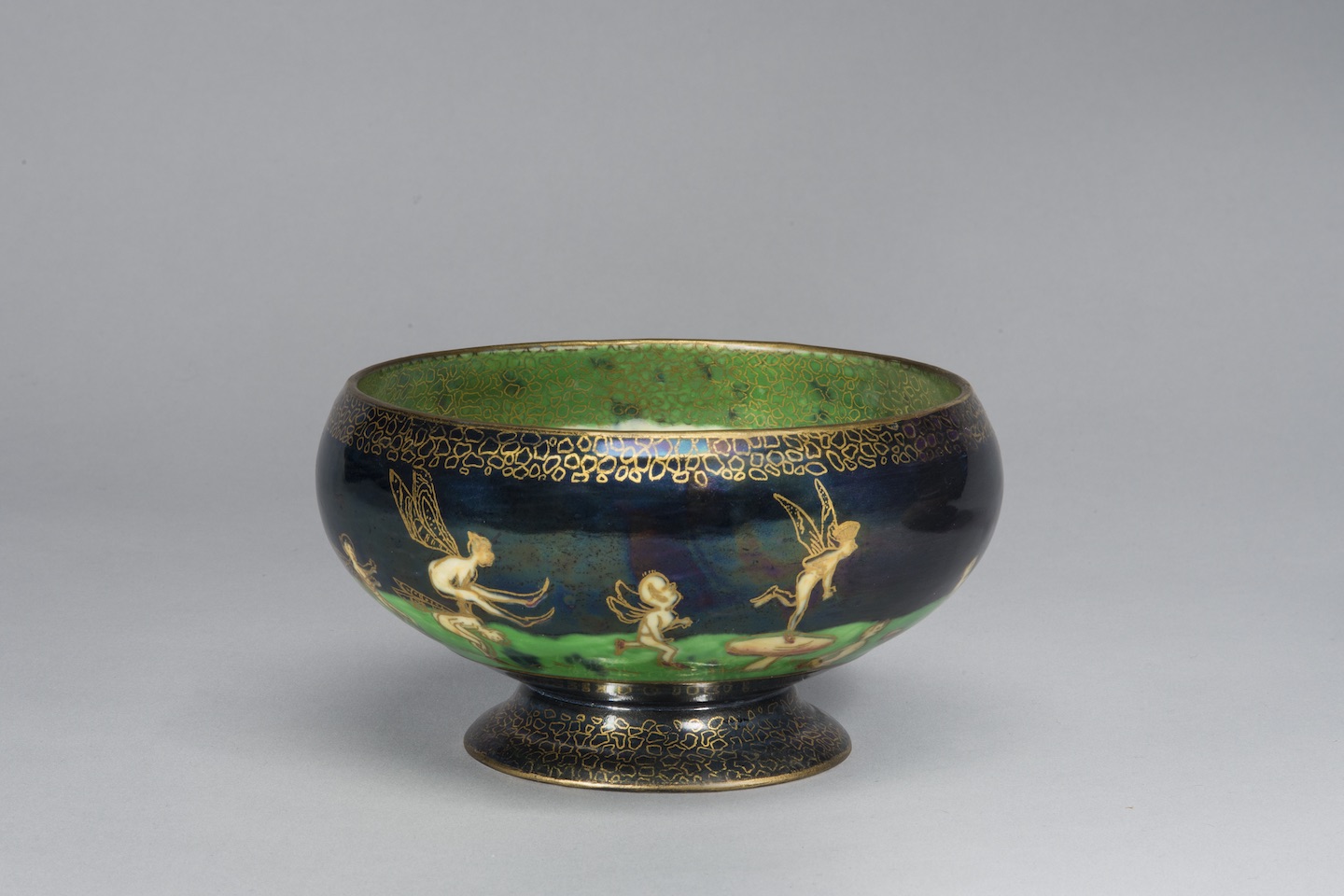 Porcelain pedestal bowl featuring an iridescent lustreware glaze. Around the bowl, winged fairies and sprites cavort on a vibrant green field set against a deep blue background. The figures and the decorative elements along the rim and foot of the bowl are outlined in gold cloisonne.