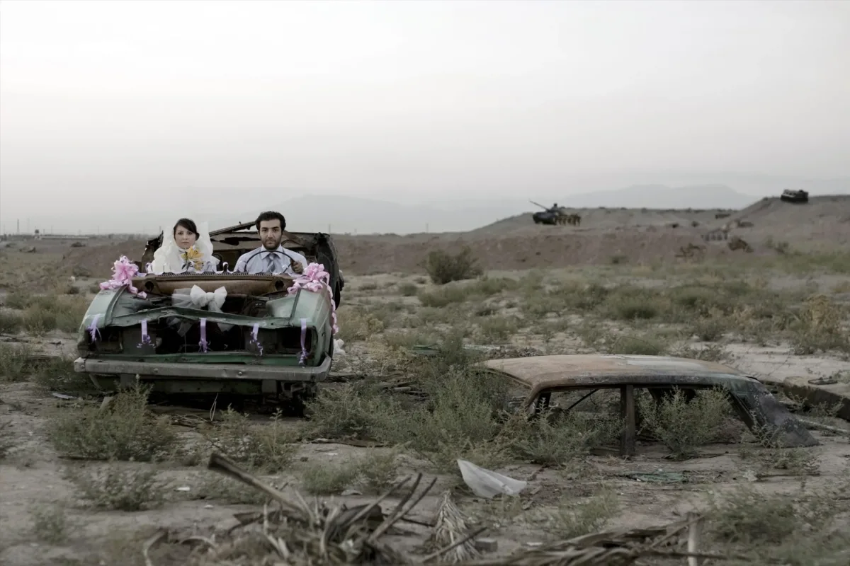 A couple sitting in a burned-out car in wedding finery, they look directly at the camera with neutral or stricken expressions.