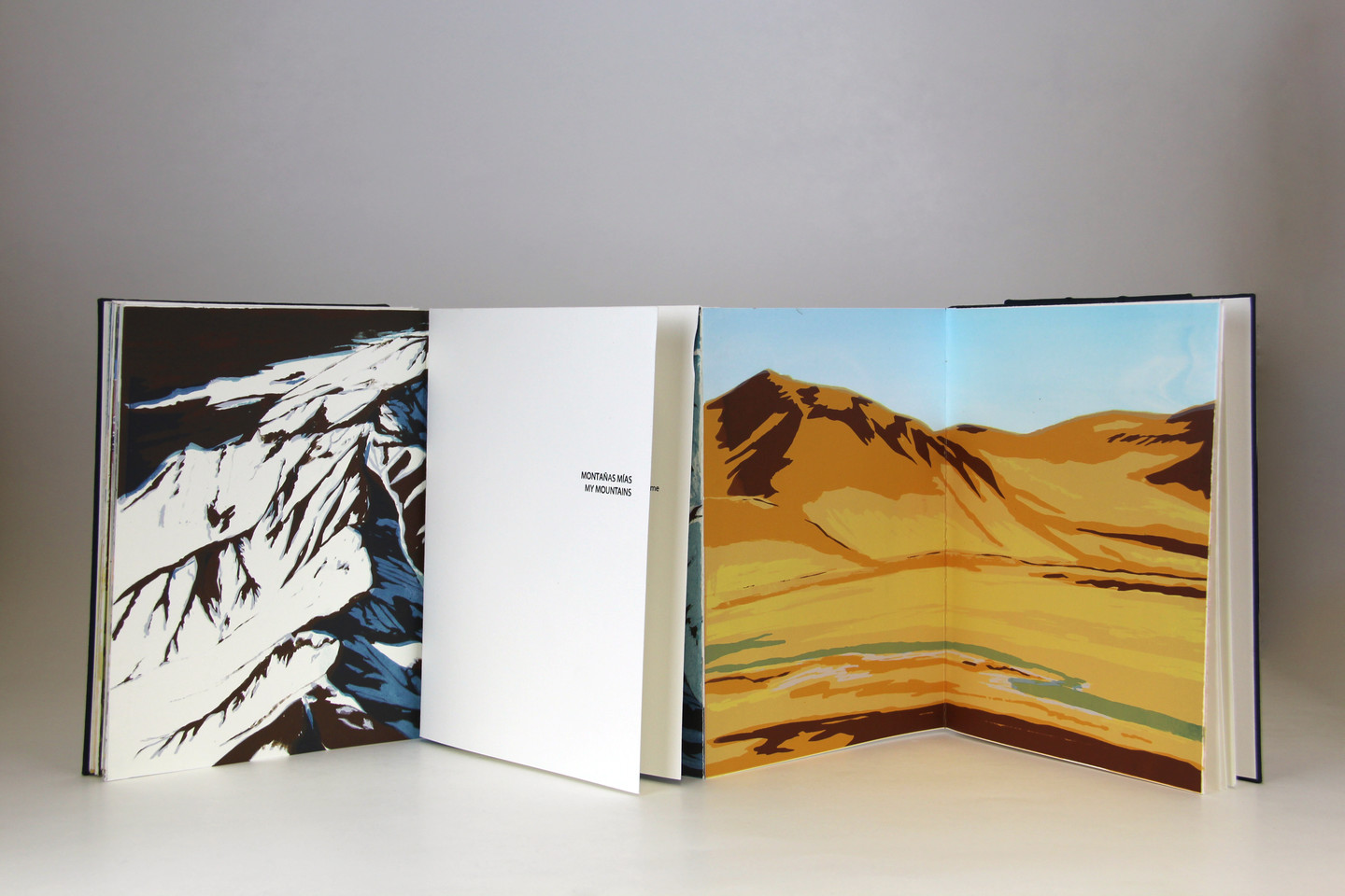 Four leaves of an open book depict mountain ranges. One image shows snow-capped mountains in blue, black and white; the other image depicts a low mountain in desert colors of beige, brown and yellow with a stream in the foreground.
