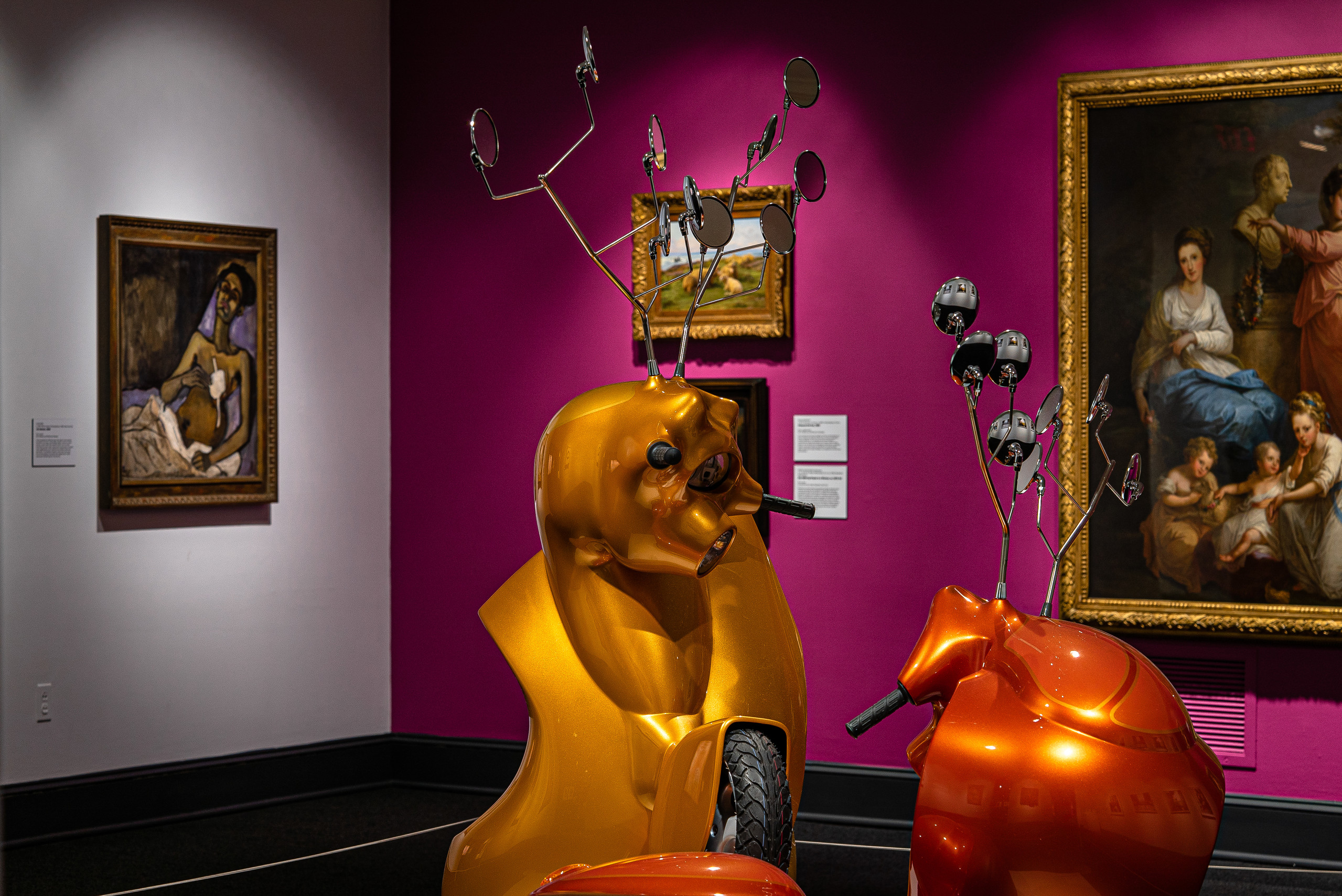 Gallery view with contemporary sculpture made of motorscooters that resemble two antlered animals fighting in foreground.Three paintings hang on a magenta wall in background. To the left on a white wall hangs another painting.