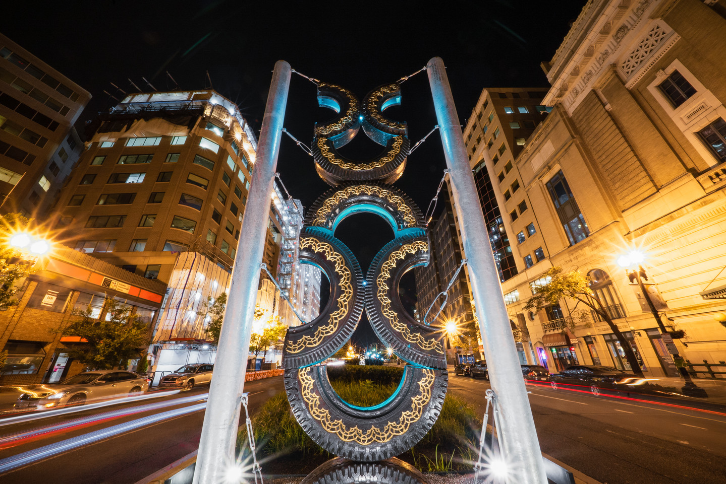 Night view of city street shows a large sculpture made of eight half-tires arranged into stacked hourglass shapes. Engravings, gold leaf, and green light on the interior of the tires decorate the structure. Light trails from cars frame the sculpture on both sides.
