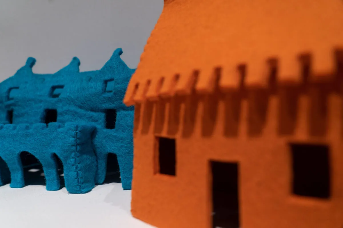 Close-up detail of a larger artwork features a view of two felt houses. On the viewer's right is an orange house that is out-of-focus while on the left is a blue house that is in-focus and has visible stitches, coarse texture, and rectangular windows.