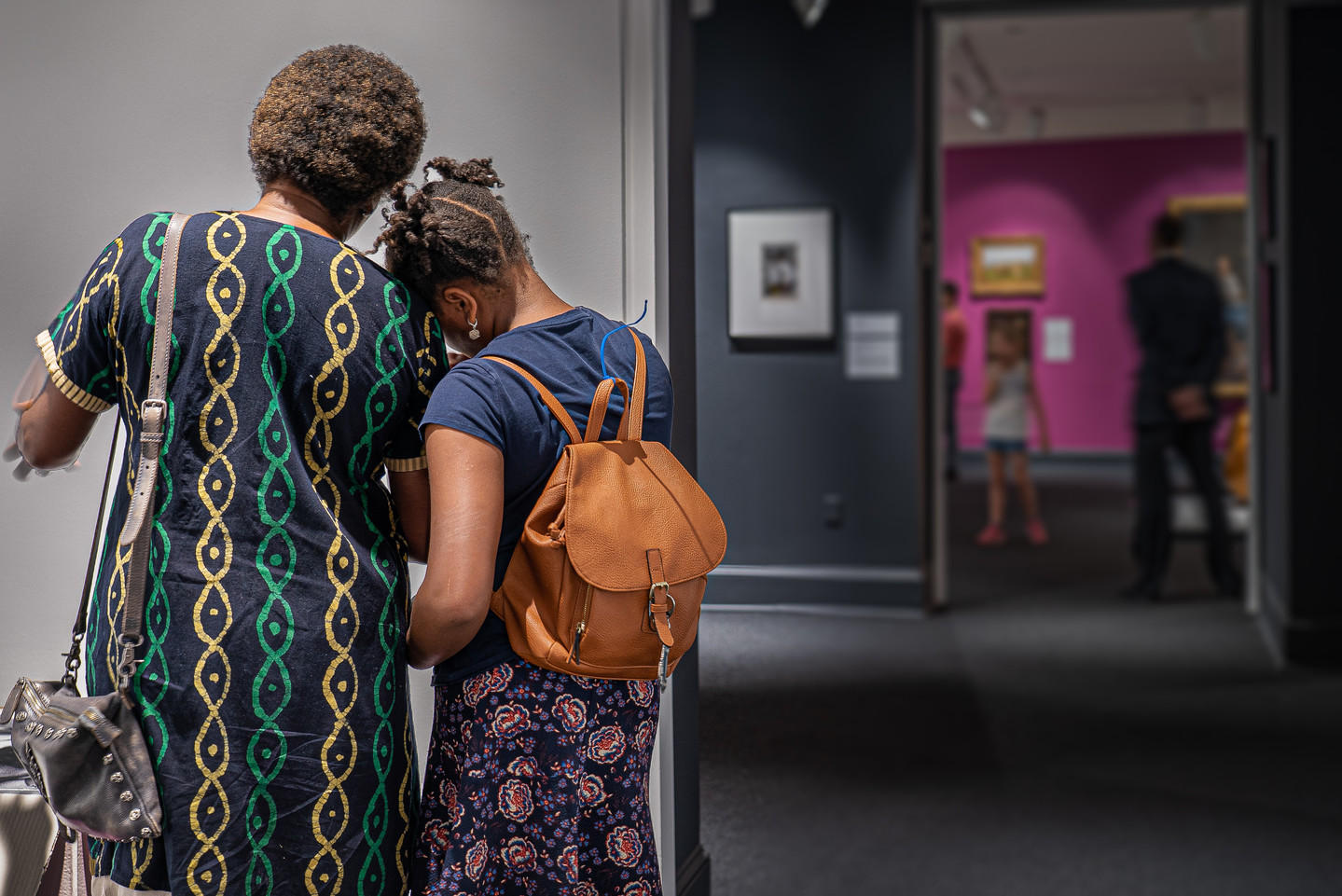A middle aged, medium dark-skinned woman and a school age medium dark skinned girl face away from the camera and lean into each other affectionately while viewing artwork.