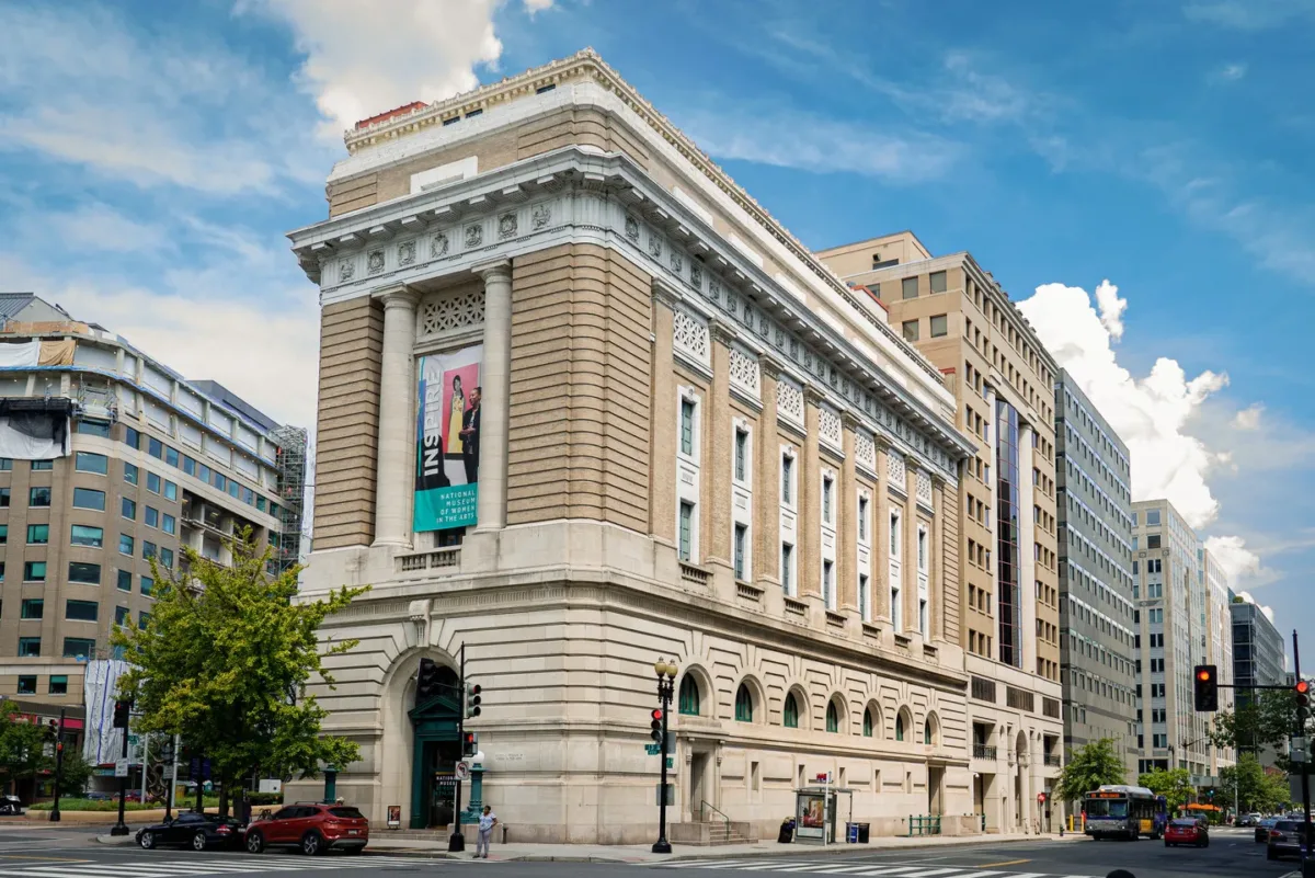 View of the museum from outside showing the Neoclassical building from one corner. The building is a tan-colored stone with an arched doorway, long vertical windows, and detailed molding around the roof.