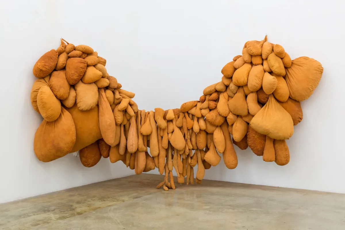 Large-scale sculpture suspended on a white wall from both ends features multiple fabric pouches in various shapes and sizes resembling proliferating biological growths.