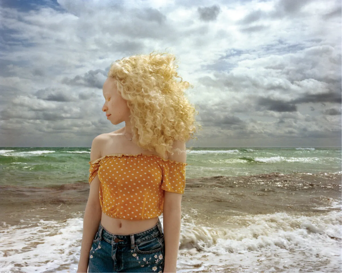 In front of a stormy ocean, a woman with light skin and blonde, wind-blown hair stands with head turned in profile and eyes closed.