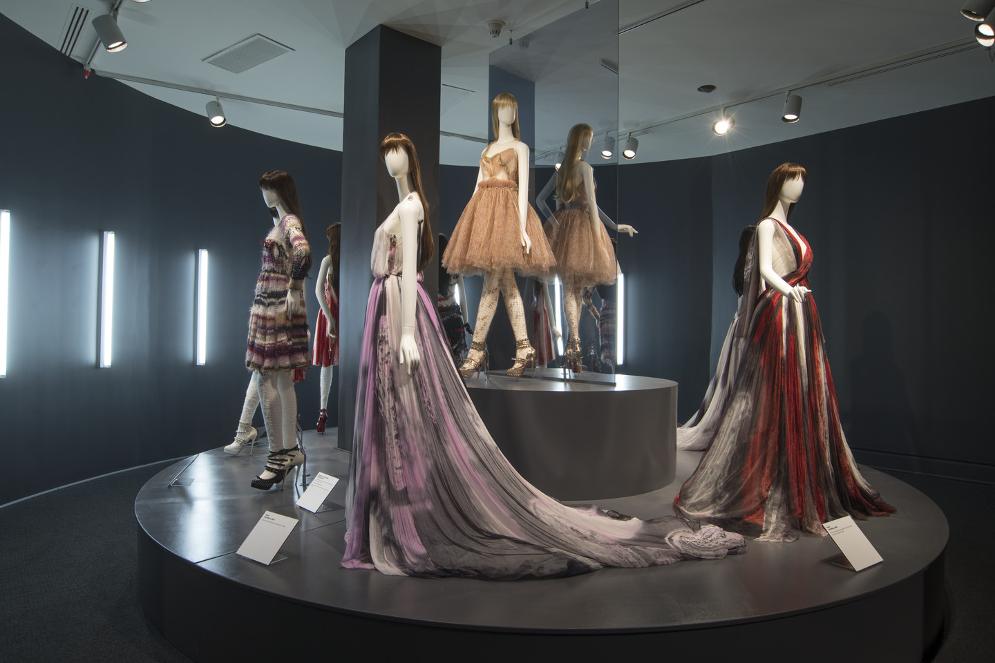 Four light skin mannequins display fashionable gowns of varying length on a circular platform.