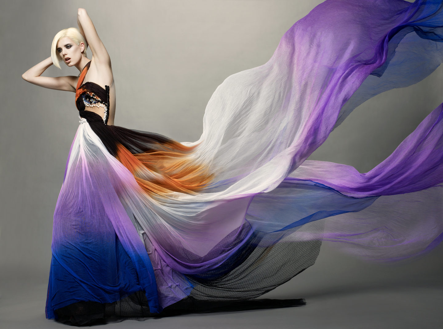 Model with light skin and platinum blond hair poses with hands behind her head wearing a long dress with shades of blue, purple, and orange. Train of dress dramatically flows off the right and top edge of the frame.