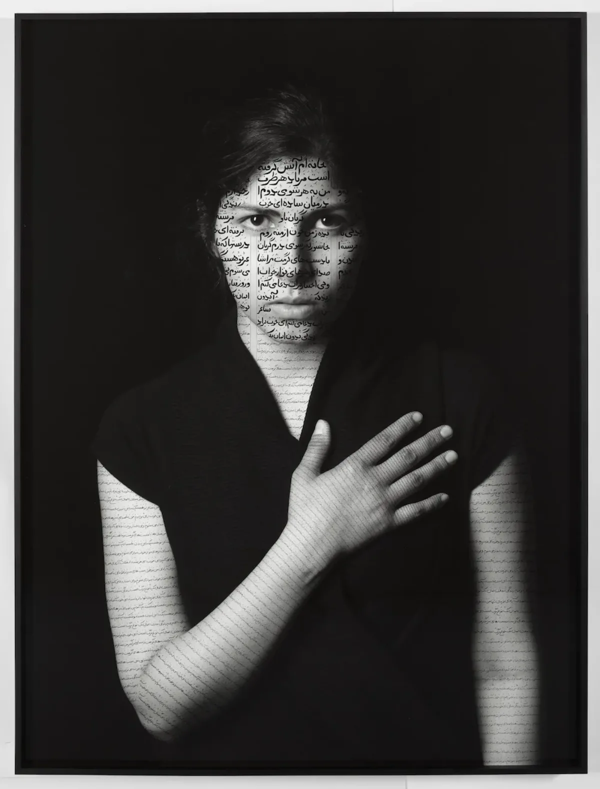 Black-and-white photograph of a woman with light skin and dark hair wearing a black t-shirt that blends into the dark black background. She holds her right hand over her heart and stares out at the viewer. Her face and arms are inscribed with calligraphic verses in Farsi.