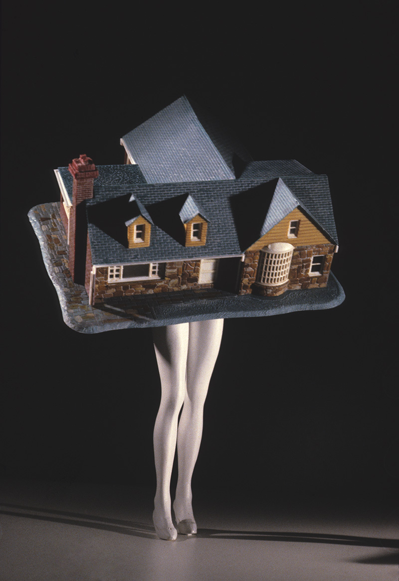 Sculpture of a large, brown brick house with black roof sits atop white mannequin legs forming a half-house, half-woman creature.