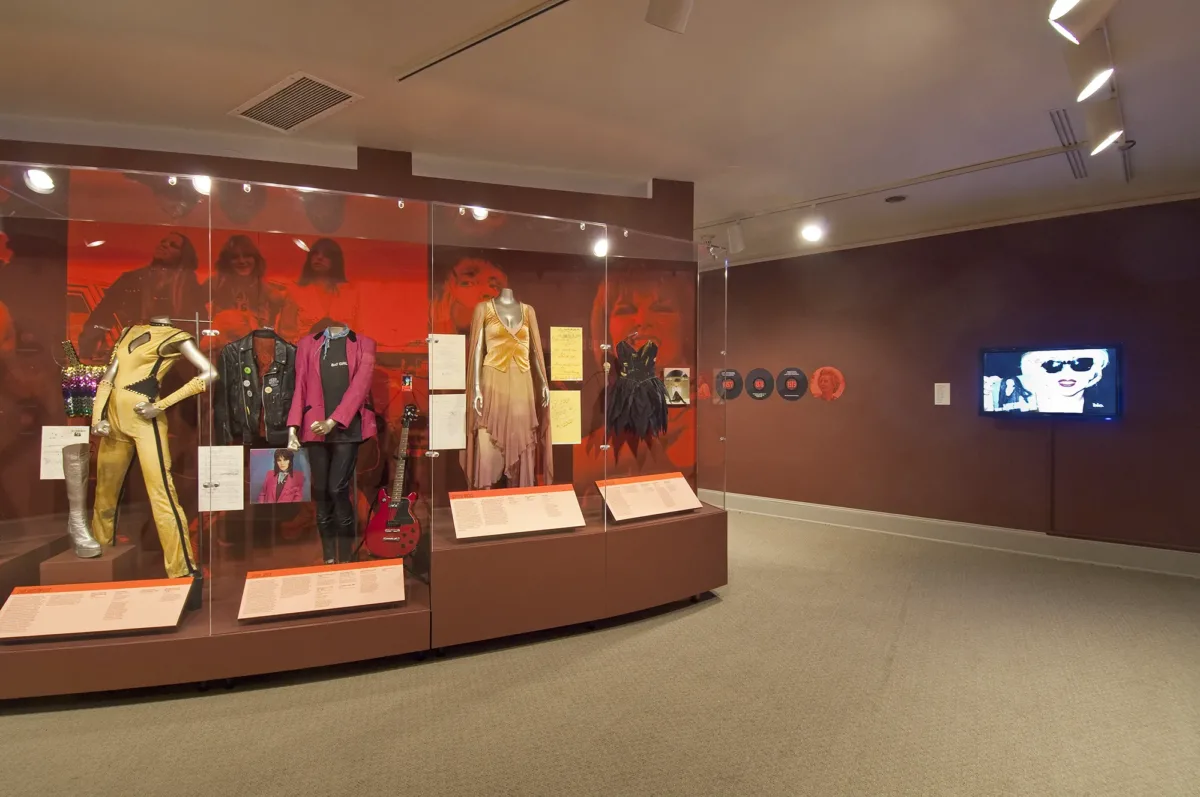 Gallery view showing a display case with a red background, its full of musician costumes.