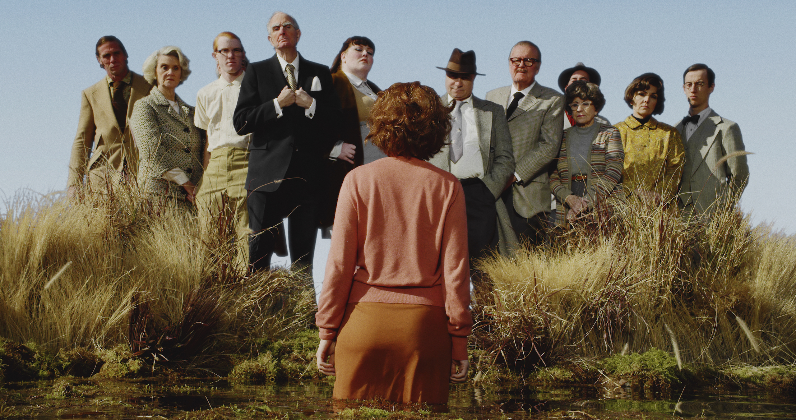 A color photograph of a woman with short red hair and pink cardigan, standing with her back to the camera in a field of tall yellow grass. She faces twelve men and women, who stand slightly elevated above her in a line and look pointedly at her.