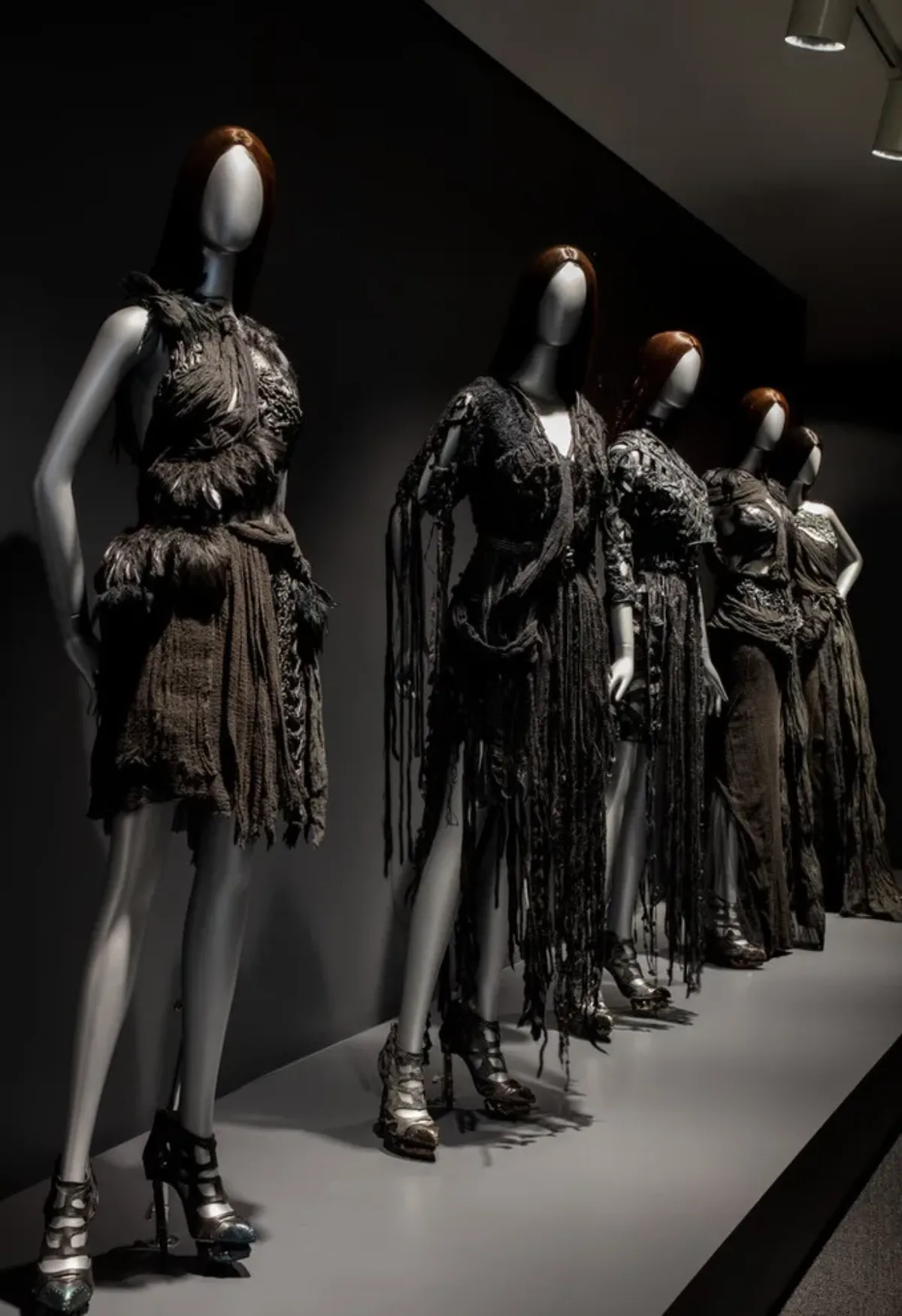 Dramatically lit installation view of Rodarte exhibition features five silver mannequins with straight brown hair wearing black dresses.