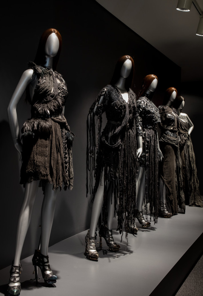 Dramatically lit installation view of Rodarte exhibition features five silver mannequins with straight brown hair wearing black dresses.