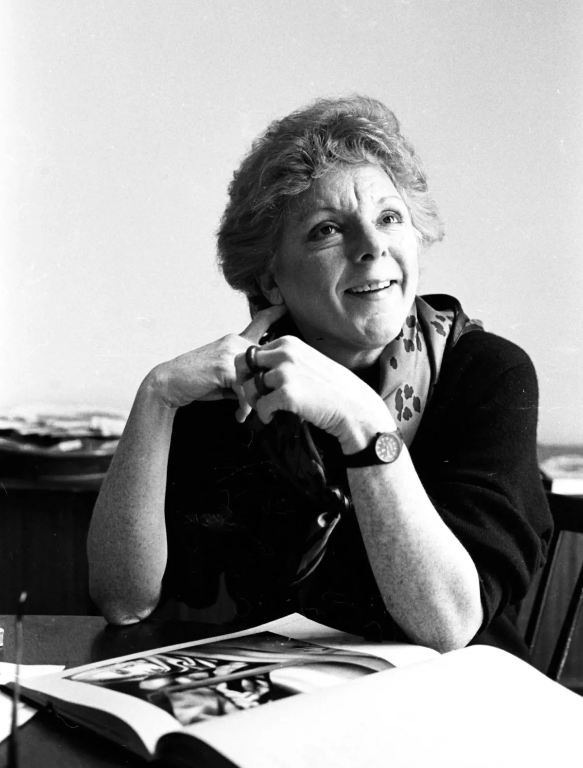 A black and white photographic portrait of seated art scholar Linda Nochlin. Dressed in a dark sweater and a print scarf, she gazes with a smile up to her left. An oversized art book is opened on the surface in front of her.