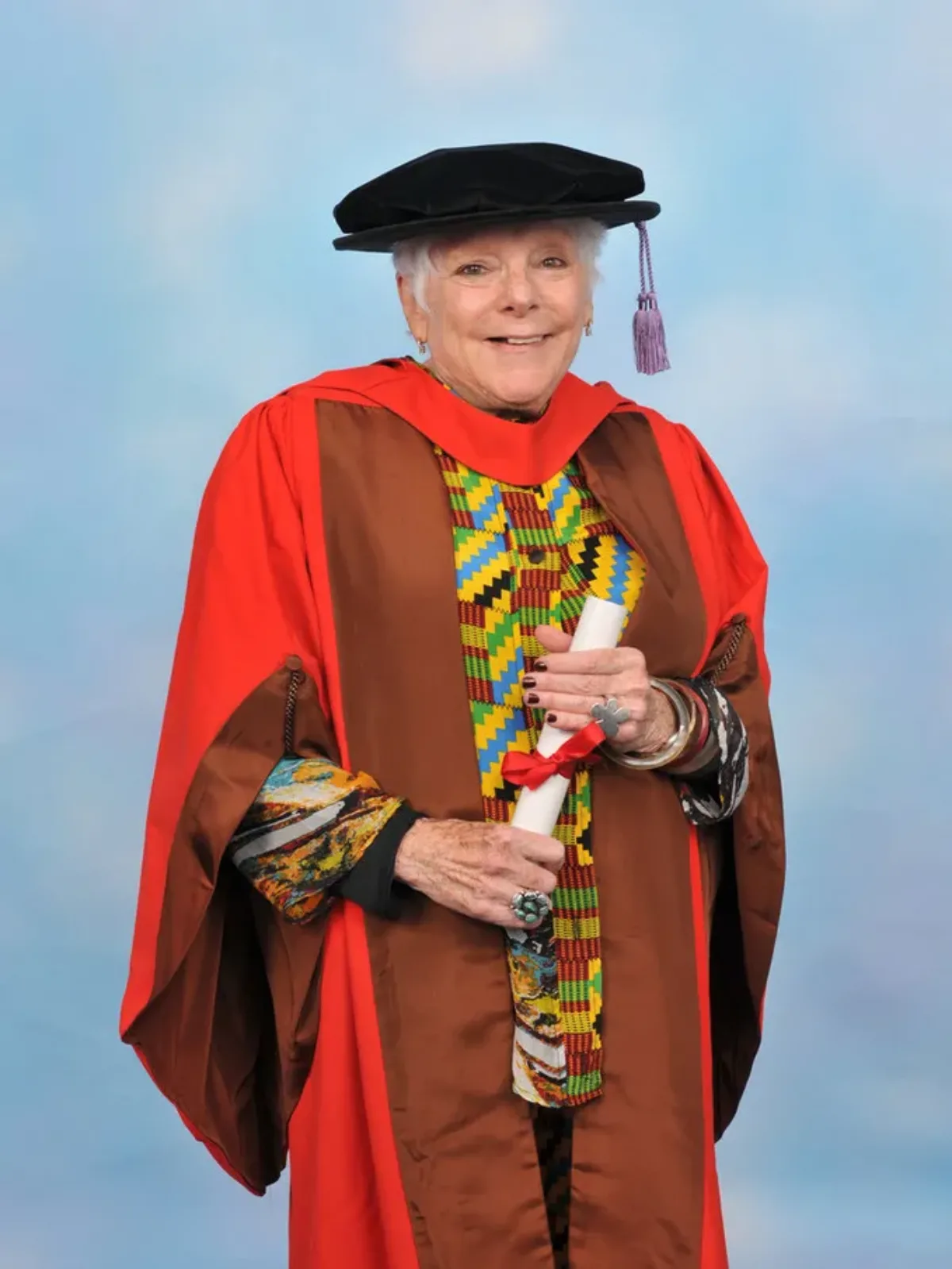 A photographic portrait of smiling art scholar Linda Nochlin in academic regalia. She wears a multicolored blouse and a black cap with a lavender tassel. Over the blouse she wears a bright red and brown gown. She holds a rolled white paper tied with a red ribbon.