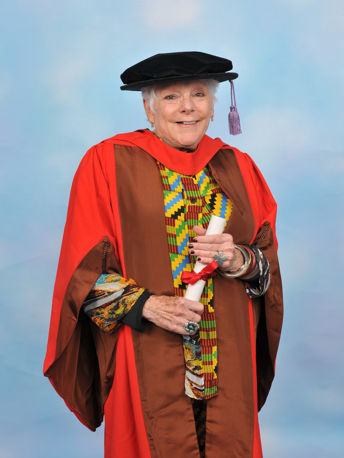 A photographic portrait of smiling art scholar Linda Nochlin in academic regalia. She wears a multicolored blouse and a black cap with a lavender tassel. Over the blouse she wears a bright red and brown gown. She holds a rolled white paper tied with a red ribbon.