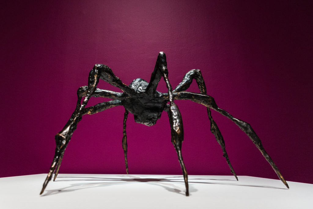 Bronze sculpture of a spider on a white platform against a magenta background.